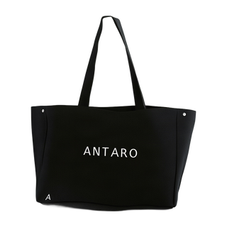 A black canvas tote bag with 'ANTARO' printed in white on the front, featuring dual shoulder straps and small silver rivets on the sides, set against a plain white background.