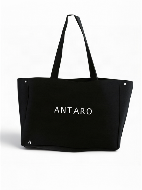 A black canvas tote bag with 'ANTARO' printed in white on the front, featuring dual shoulder straps and small silver rivets on the sides, set against a plain white background.